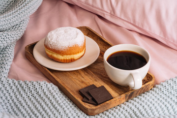 Morning breakfast in bed with aromatic and hot coffee, a doughnut in powdered sugar and three slices of chocolate on a wooden tray against a backdrop of pink sheets, pillows and blue plaid