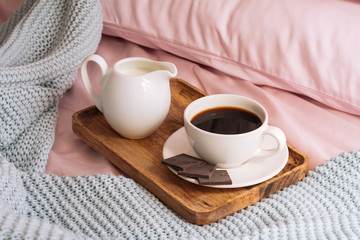 Fototapeta na wymiar Breakfast in bed with freshly brewed and delicious coffee, a jug of cold milk and three slices of real bittersweet chocolate on a wooden tray against a backdrop of pink bedding and a blue plaid