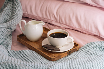 Fototapeta na wymiar Breakfast in bed with freshly brewed and delicious coffee, a jug of cold milk and three slices of real dark chocolate on a wooden tray against a backdrop of pink sheets, pillows and a blue plaid