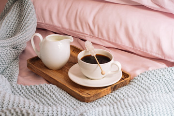 Fototapeta na wymiar Breakfast in bed with freshly brewed and delicious coffee, a jug of cold milk and crystal sugar on a stick on a wooden tray against a backdrop of pink sheets, pillows and a blue plaid
