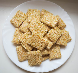 In a white round plate are square-shaped cookies of white sesame, honey and gluten-free sugar on a light table