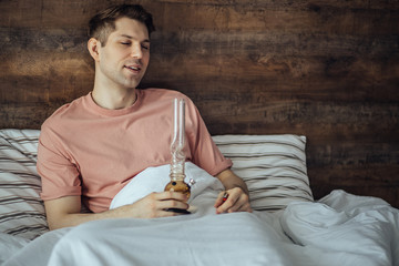 young man smokes bong cannabis weed at home. medical marijuana, hemp use and legalization of the...