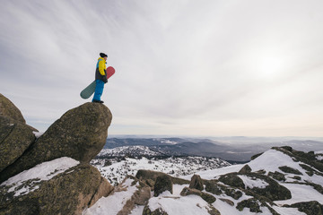Hiker snowboarder freerider holding snowboard standing on big rock cliff watching mountain landscape