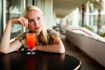A beautiful young woman of European appearance is drinking an exotic summer cocktail while sitting at a table of a street summer cafe.