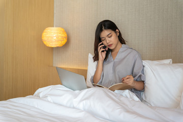 Young asian woman relaxing in the hotel and working with laptop on a bed, calling to cusotmer, Business trip, Working travel concept
