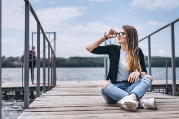 Young woman with long hair in stylish glasses posing on a wooden pier near the lake. Girl dressed in jeans and t-shirt smiling and looking at the camera