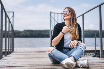 Young woman with long hair in stylish glasses posing on a wooden pier near the lake. Girl dressed in jeans and t-shirt smiling and looking at the camera