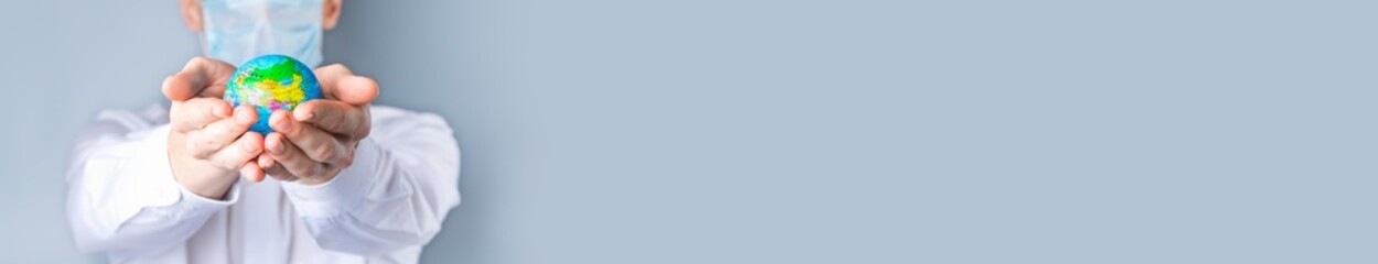 Close-up of a man's hands holding a small globe, a man in a medical mask out of focus, dressed in a white shirt, uniform on a gray background.  Banner. 2020, the global economic crisis. Coronavirus