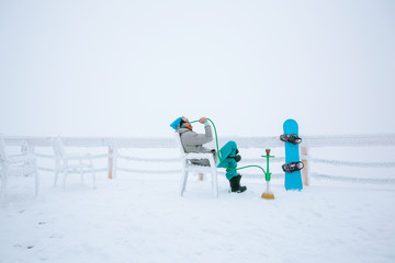 Snowboarder man relaxing and smoke a hookah or shisha, enjoying mountain view, winter frozen foggy day, snow and frost on the branch and table outdoor