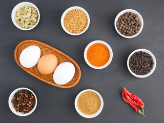 Raw chicken eggs, black pepper, turmeric, masala powder, red chili, cloves, allspice, green cardamom on black wooden background. Ingredients for making scrambled eggs or omelet. Organic food concept