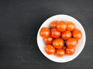 Red small cherry tomato in white bowl on black wooden background with copy space. Healthy eating and organic food concept