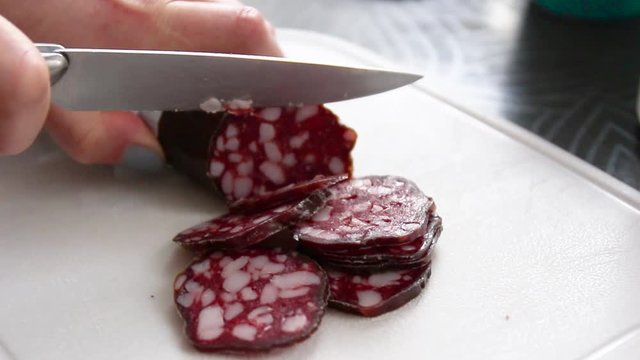 Person hands with a knife cutting sausage salami on a cutting board in the kitchen.