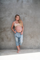 Happy pretty pregnant woman in denim jumpsuit and bra standing over gray wall background. Maternity concept