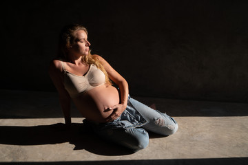 Fototapeta na wymiar Pretty pregnant woman in denim jumpsuit and bra sitting on the concrete floor in sunlight. Maternity concept