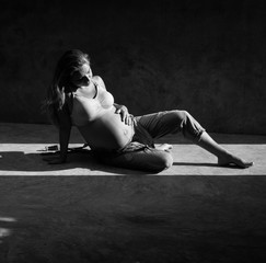 Pretty pregnant woman in denim jumpsuit and bra sitting on the concrete floor in sunlight. Maternity concept
