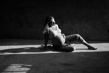 Pretty pregnant woman in denim jumpsuit and bra sitting on the concrete floor in sunlight. Maternity concept