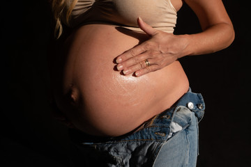 Cropped image of pregnant woman in denim jumpsuit and bra applying lotion on her belly. Maternity, pregnancy, new life and health care concept