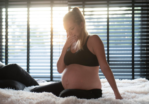 Pretty pregnant woman in black leggins and bra covering mouth with hand while sitting on her bed at home. Maternity, health care concept