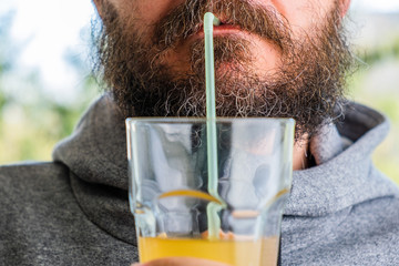 Bearded man in hoodie drinking juice with straw outdoor, hands and lips detail