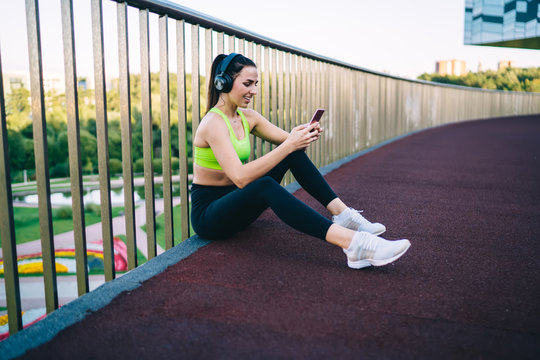 Positive caucasian sportswoman meloman checking playlist songs via app on mobile phone sitting outdoors, smiling female fitness blogger share media content with followers enjoying chat via smartphone