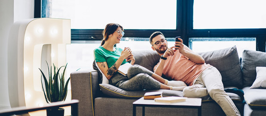 Cheerful couple in love having fun at home interior joking and share funny photos on mobile phones via bluetooth connection, smiling young marriage watching online videos on cellulars together