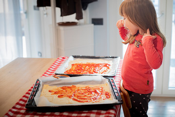 Child girl in a kitchen tasting some tomato sauce from raw pizza before baking. Lockdown activity idea for kids.