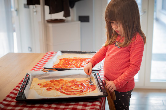 Child girl standing on a chair spreading tomato sauce on raw pizza before baking. Lockdown activity idea for kids. Side view.