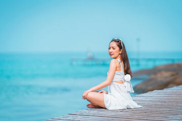 Fototapeta na wymiar girl in white dress and sunglasses a posing sit relax on the walkway bridge wood beach with sunlight shine and blue sky