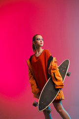 A young girl with bright makeup holds a longboard in her hands on a fashionable neon color background. Portrait of a girl skater