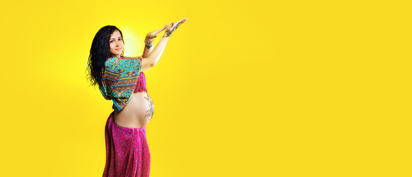 Pregnant happy woman in Indian sari with mehendi tattoo painted with henna on belly, fun fooling over yellow background, panoramic mock-up with space for text