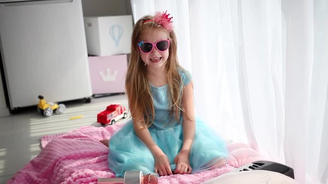 Little girl in a beautiful blue dress, a crown and sunglasses laughs looking at the camera.  Stay at home