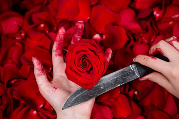 Close-up of hands in blood holding a knife and a rose bud, against a background of roses and petals.