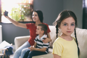 Unhappy little girl feeling jealous while mother taking selfie with her brother at home