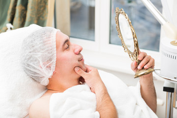 Man examines face in mirror lying in beauty salon