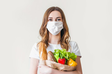 Young woman in medical mask holds an eco paper bag with food, fruits and vegetables, pepper, baguette, lettuce, safe online smart delivery, coronovirus, quarantine, pandemic, stay home concept