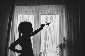 Silhouette of a happy child girl playing with a toy airplane in her room by the window. Dream to fly. Close the borders of air travel.