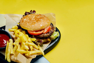 Appetizing American burger with beef, tomatoes, lettuce and fries on a black tray on a colored background. Tasty fast food