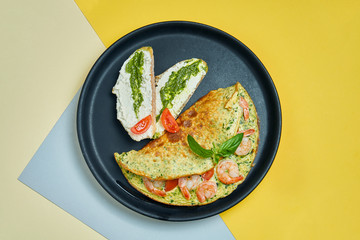Continental breakfast - omelet with shrimps, tomatoes and basil with toast with cream cheese and pesto on a black plate on colored backgrounds.
