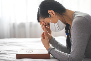 Woman with mask praying next to bed with her bible near.