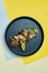 Close up view on Appetizing toast on rye bread with arugula, poached egg, salmon and sun-dried tomatoes on a black plate on colored background. Breakfast food