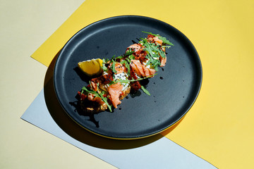 Close up view on Appetizing toast on rye bread with arugula, poached egg, salmon and sun-dried tomatoes on a black plate on colored background. Breakfast food