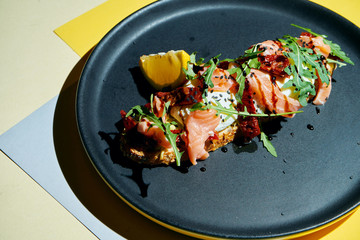 Close up view on Appetizing toast on rye bread with arugula, poached egg, salmon and sun-dried tomatoes on a black plate on colored background. Breakfast food