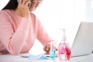 Business woman hand using smartphone with work from home. Laptop mouse, sanitizer gel, face mask, and alcohol spray for prevention Coronavirus disease (COVID-19). Healthcare disinfection concept.