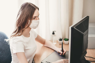 Young girl artist designer freelancer in a medical mask sits at home at the table and draws on a...