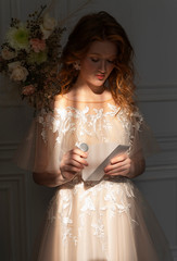 Beautiful natural redhead girl bride, with nude makeup, wearing a white dress, poses in the shadow, illuminated by a narrow beam of light from the window, opens the letter envelope.