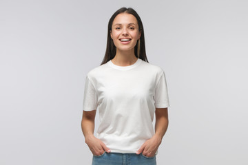 Laughing student girl wearing white T-shirt and blue jeans, standing with hands in pockets, looking...