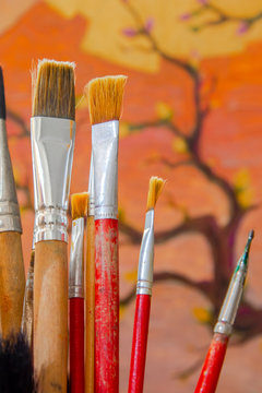 Some artistic paintbrushes in front of an unfinished painting.