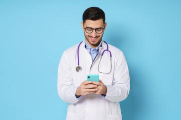 Fototapeta premium Young male doctor in white coat smiling while looking at screen of his phone, using medical app, standing isolated on blue background