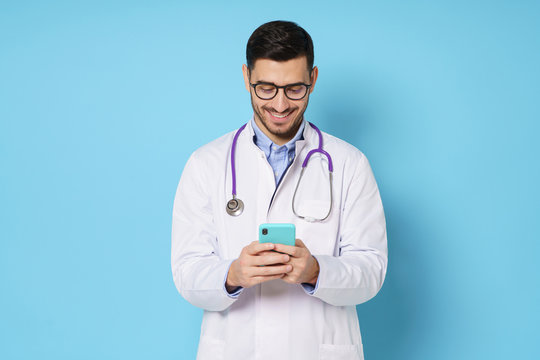 Young male doctor in white coat smiling while looking at screen of his phone, using medical app, standing isolated on blue background