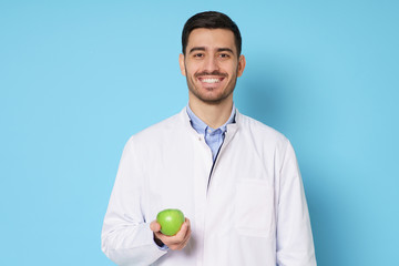 Smiling nutritionist or dentist doctor wearing white coat and round glasses, holding green apple in hand, isolated on blue background